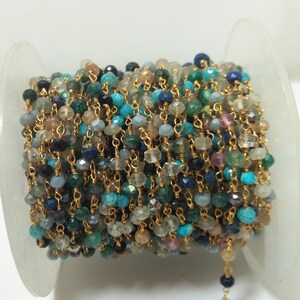 Op de afbeelding: Een spoel met een gouden ketting met verschillende gekleurde edelsteen kralen, waaronder turquoise, blauw, groen en zwart. De ketting is om een witte spoel gewikkeld.