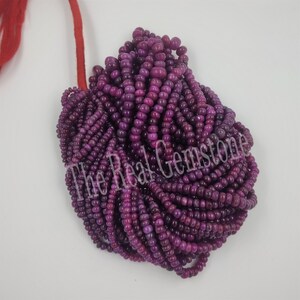 May include: A large bundle of deep purple gemstone beads. The beads are small, cylindrical, and strung together. A red string is visible at the top. The text "The Real Gemstone" is printed across the beads.