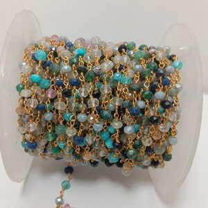 Op de afbeelding: Een spoel met een gouden ketting met verschillende gekleurde edelsteen kralen, waaronder blauw, groen en bruin. De ketting is om een witte spoel gewikkeld.