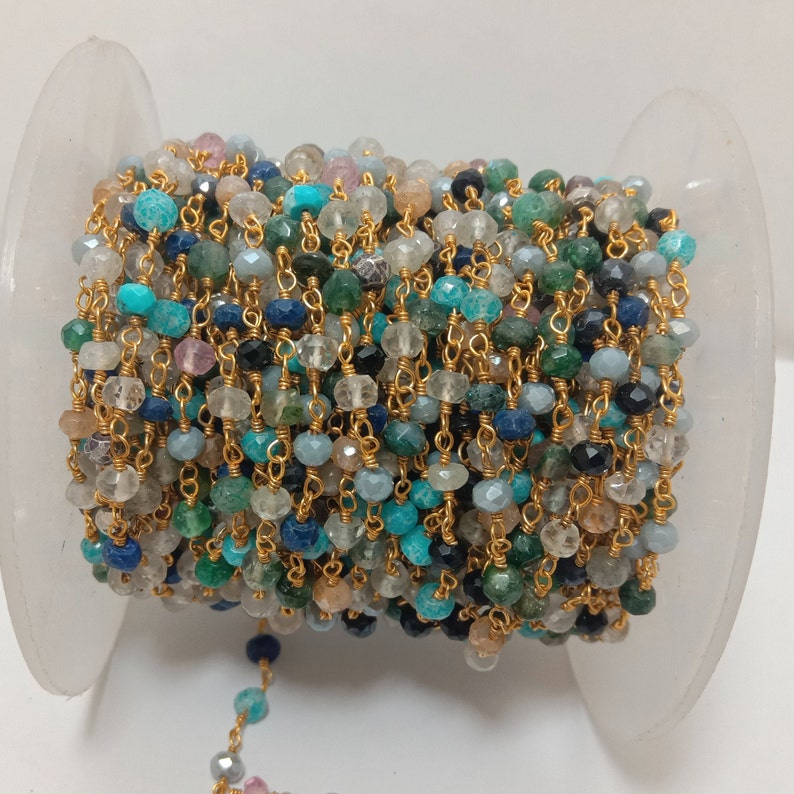 Op de afbeelding: Een spoel met een gouden ketting met verschillende gekleurde edelsteen kralen, waaronder blauw, groen en bruin. De ketting is om een witte spoel gewikkeld.