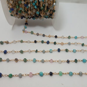 Op de afbeelding: Een gouden ketting met verschillende gekleurde edelsteen kralen, waaronder blauw, groen en roze. De ketting ligt in rijen op een wit oppervlak.