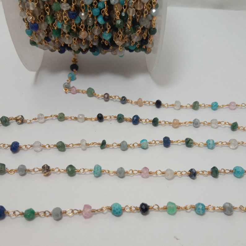 Op de afbeelding: Een gouden ketting met verschillende gekleurde edelsteen kralen, waaronder blauw, groen en roze. De ketting ligt in rijen op een wit oppervlak.