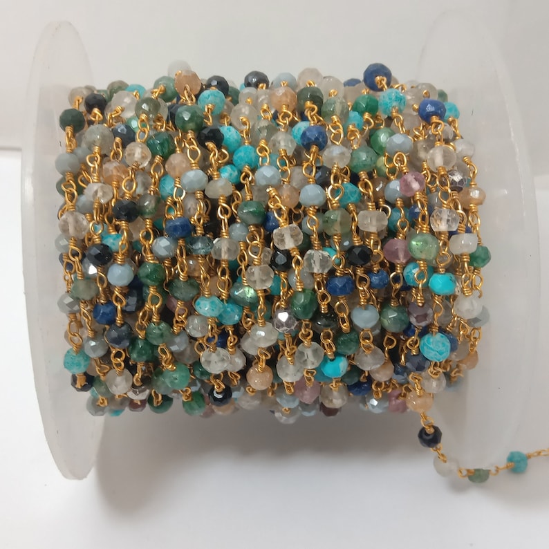 Op de afbeelding: Een gouden ketting met verschillende gekleurde edelsteen kralen, waaronder blauw, groen en bruin. De ketting is om een witte spoel gewikkeld.