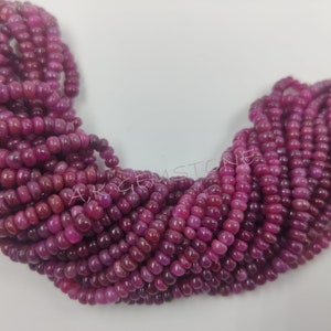 May include: A strand of deep pink ruby beads, each bead is round and slightly elongated, creating a textured look. The beads are arranged in a loose pile, showcasing their colour and shine.