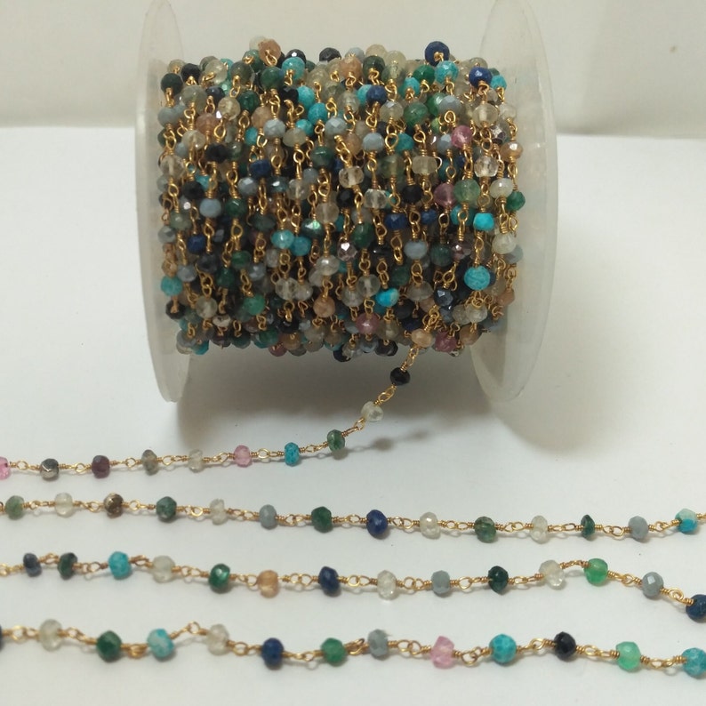 Op de afbeelding: Een spoel met een gouden ketting met kleine, kleurrijke edelsteen kralen. De ketting is gemaakt van verguld metaal en heeft een verscheidenheid aan edelstenen, waaronder turquoise, lapis lazuli en groene aventurijn.
