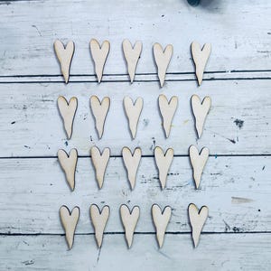 May include: Twenty wooden heart cutouts with a natural wood finish are arranged on a white and gray wooden surface. The hearts have a unique teardrop shape at the bottom. The image is well-lit, highlighting the wood grain.