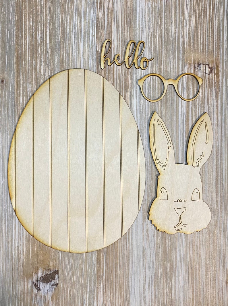 May include: Wooden Easter-themed cutouts on a light wood grain surface. Includes an egg shape with vertical lines, a bunny head, glasses, and the word "hello". The cutouts are unfinished and ready for painting or decorating.