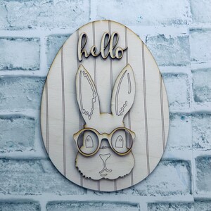 May include: Wooden Easter egg decoration with a bunny wearing glasses. The word "hello" is written above the bunny. The egg has vertical stripes and is made of light-colored wood.