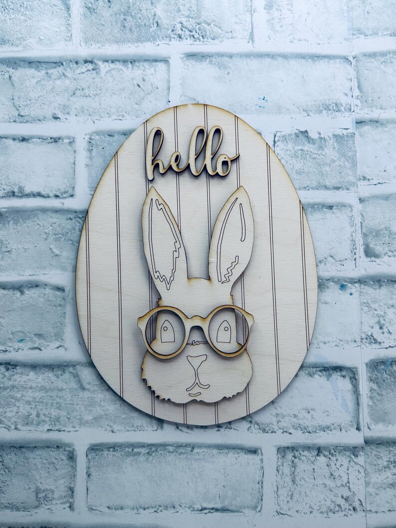 May include: Wooden Easter egg decoration with a bunny wearing glasses. The word "hello" is written above the bunny. The egg has vertical stripes and is made of light-colored wood.