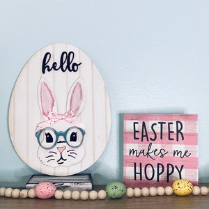 May include: Easter-themed decor featuring a wooden egg with a bunny wearing glasses and a pink bow, with the word "hello" above. A sign with the text "EASTER makes me HOPPY" is also present. A garland of wooden beads and speckled eggs completes the scene.