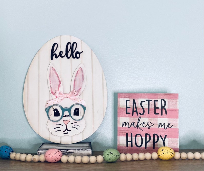 May include: Easter-themed decor featuring a wooden egg with a bunny wearing glasses and a pink bow, with the word "hello" above. A sign with the text "EASTER makes me HOPPY" is also present. A garland of wooden beads and speckled eggs completes the scene.