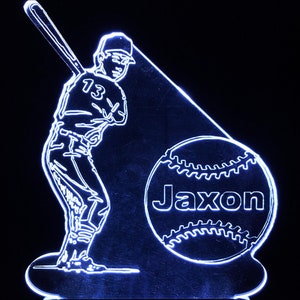 May include: A white LED light with a baseball player silhouette holding a bat and a baseball with the name "Jaxon" on it. The light is on a black background.