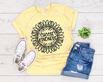 May include: A yellow t-shirt with a black sunflower graphic and the text "Choose Kindness". The shirt is laid out on a wooden surface with a pair of white sneakers and denim shorts.