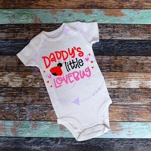 May include: White baby onesie with the text "Daddy's little lovebug" in pink and black lettering. A red ladybug with black spots is above the text. Pink hearts are scattered around the text.