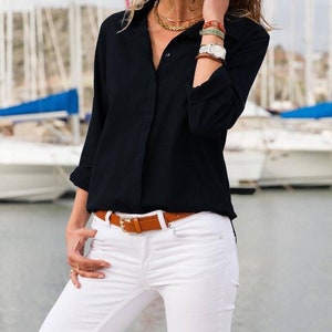 May include: A black button-down shirt with rolled-up sleeves, paired with white jeans and a brown belt. The belt has a gold-colored buckle. The outfit is accessorized with gold necklaces and bracelets. The background features boats.
