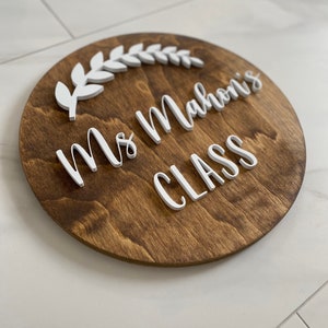 Puede incluir: Un letrero redondo de madera con el texto "Ms. Mahoni's Class" en letras blancas en relieve. Un dise&ntilde;o de hojas blancas est&aacute; por encima del texto.