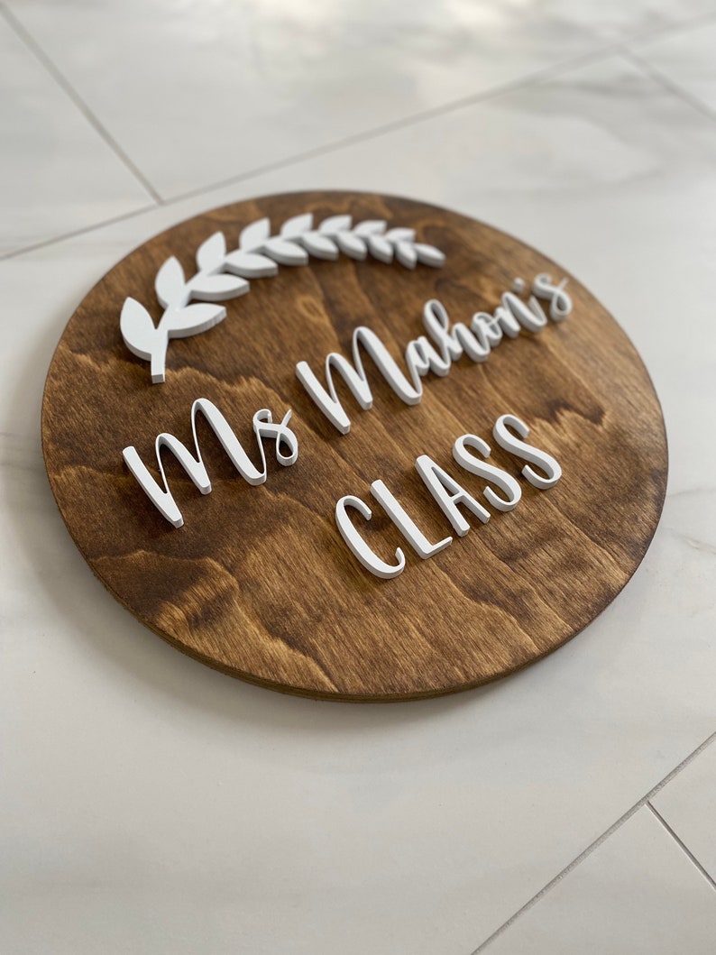Puede incluir: Un letrero redondo de madera con el texto "Ms. Mahoni's Class" en letras blancas en relieve. Un dise&ntilde;o de hojas blancas est&aacute; por encima del texto.