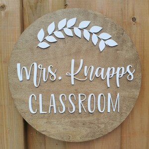 Puede incluir: Un letrero redondo de madera con una corona de hojas blancas y el texto "Mrs. Knapps Classroom" en letras blancas.