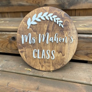 Puede incluir: Un letrero redondo de madera con una mancha marr&oacute;n. El letrero tiene letras blancas que dicen "Ms. Mahon's Class" y un dise&ntilde;o de hojas blancas.
