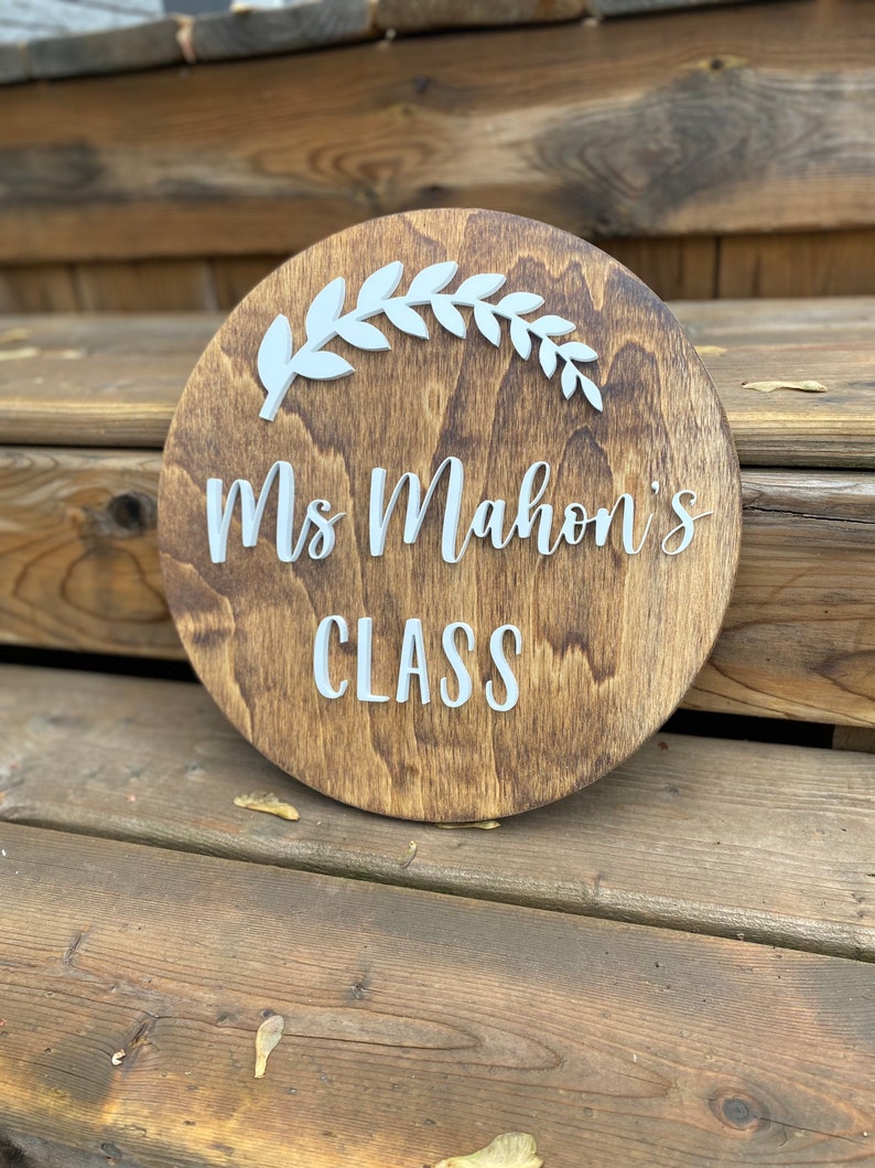 Puede incluir: Un letrero redondo de madera con una mancha marr&oacute;n. El letrero tiene letras blancas que dicen "Ms. Mahon's Class" y un dise&ntilde;o de hojas blancas.