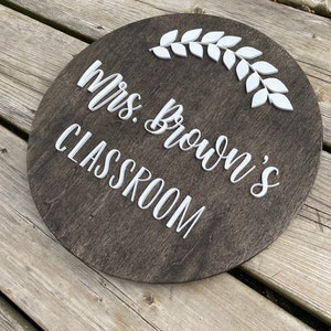 Puede incluir: Un letrero redondo de madera marr&oacute;n oscuro con letras blancas que dice "Mrs. Brown's Classroom" con un dise&ntilde;o de rama de hojas blancas.