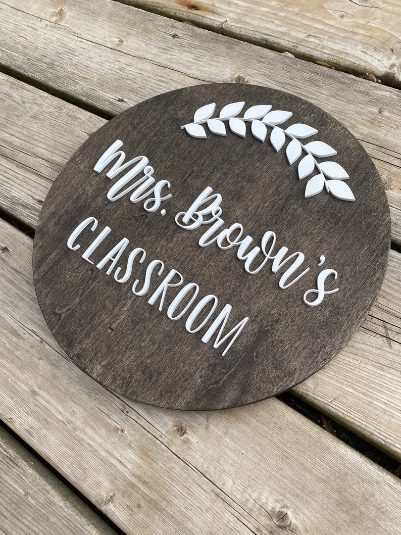 Puede incluir: Un letrero redondo de madera marr&oacute;n oscuro con letras blancas que dice "Mrs. Brown's Classroom" con un dise&ntilde;o de rama de hojas blancas.