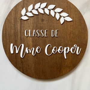 Puede incluir: Un letrero redondo de madera con una corona blanca y el texto "CLASSE DE Mme Cooper" en letras blancas.