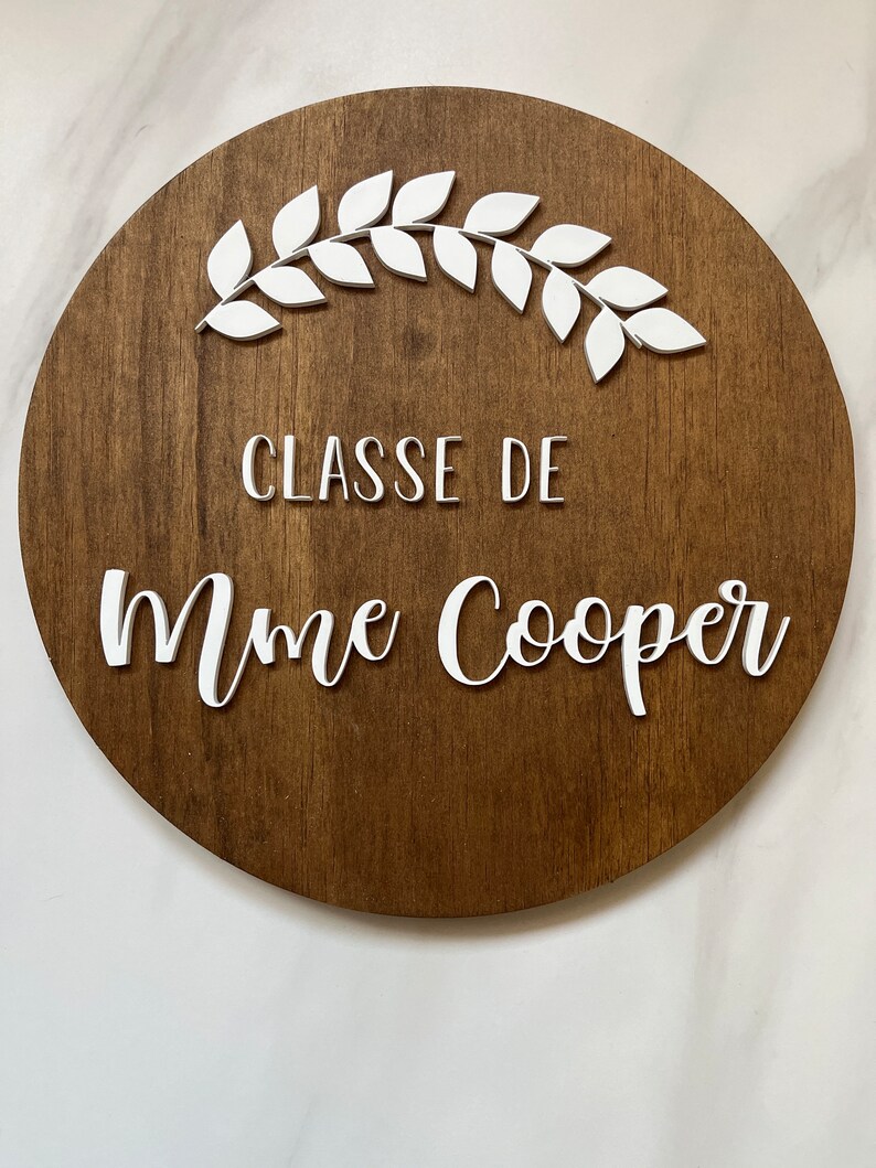 Puede incluir: Un letrero redondo de madera con una corona blanca y el texto "CLASSE DE Mme Cooper" en letras blancas.
