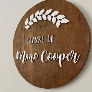 Puede incluir: Un letrero redondo de madera con una corona blanca pintada y el texto "CLASSE DE Mme Cooper" en blanco.