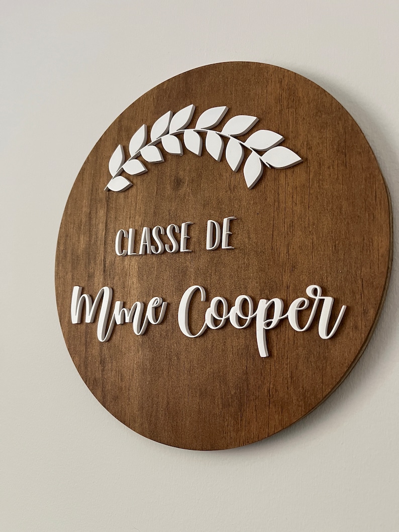 Puede incluir: Un letrero redondo de madera con una corona blanca pintada y el texto "CLASSE DE Mme Cooper" en blanco.