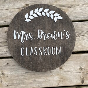 Puede incluir: Un letrero redondo de madera con una mancha marr&oacute;n oscura. El letrero tiene letras blancas que dicen "Mrs. Brown's Classroom" y una guirnalda de hojas blancas.