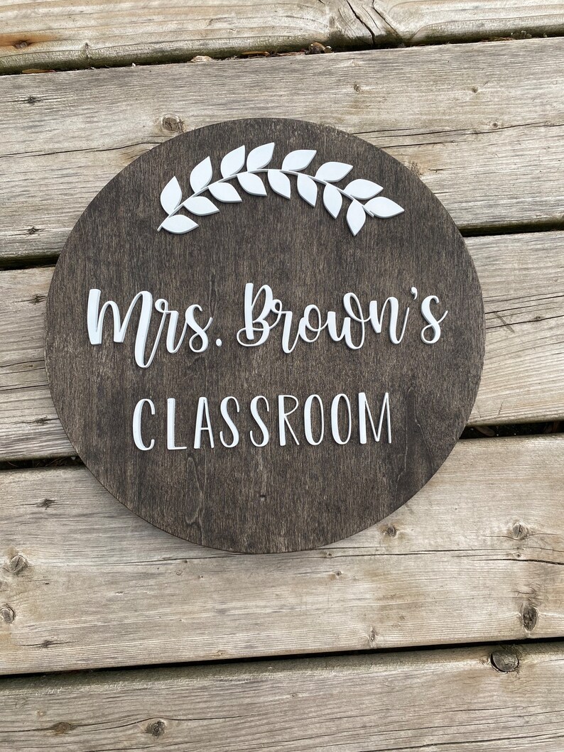 Puede incluir: Un letrero redondo de madera con una mancha marr&oacute;n oscura. El letrero tiene letras blancas que dicen "Mrs. Brown's Classroom" y una guirnalda de hojas blancas.