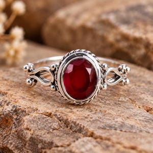 May include: A silver ring featuring an oval-cut deep red gemstone set in a twisted rope-style bezel. The band displays intricate metalwork with small spherical accents and openwork detailing on both sides of the central stone setting.