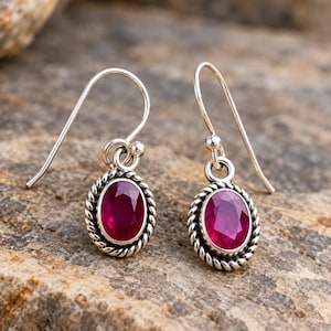 May include: A pair of silver drop earrings featuring oval-shaped, faceted deep pink stones. Each stone is set within a twisted rope-style silver border and hangs from a simple silver hook wire, creating a classic and balanced jewellery design.