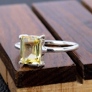May include: A silver ring with a yellow gemstone set in an emerald cut. The ring is on a wooden surface.