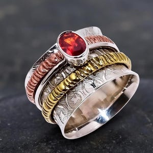 Op de afbeelding: Een brede zilveren spinnerring met een centrale ovaal geslepen rode edelsteen. De ring bevat een getextureerde koperkleurige draad, een gedraaide goudkleurige draad en een centraal zilveren gedeelte met een ingewikkeld gegraveerd bloemmotief.
