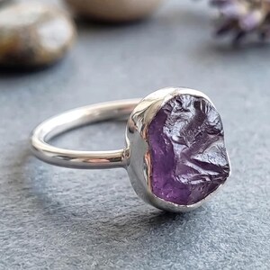 May include: A silver ring featuring a single, raw, uncut purple amethyst gemstone set in a smooth metal bezel. The band is polished and rounded, showcasing a natural, irregular texture on the surface of the deep violet stone.