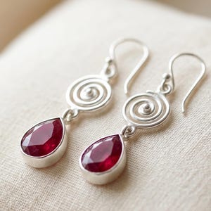 Könnte beinhalten: Silberne Ohrringe mit einem Spiralmuster und tropfenförmigen, tiefroten Edelsteinen. Die Ohrringe werden auf cremefarbenem Stoff präsentiert, wodurch das elegante Design und die Farbe des Schmucks hervorgehoben werden.
