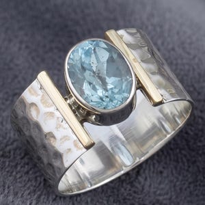 May include: A silver ring with a hammered texture and gold detailing. The ring showcases a large, oval-cut, light blue gemstone. The band has a wide design with gold bars either side of the gemstone.