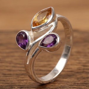 Könnte beinhalten: Ein silberner Ring mit drei Edelsteinen. Er hat einen gelben Edelstein im Marquiseschliff und zwei birnenförmige violette Edelsteine. Der Ring hat ein florales Design, wobei das Band elegant geschwungen ist. Die Edelsteine sind in Silber gefasst.