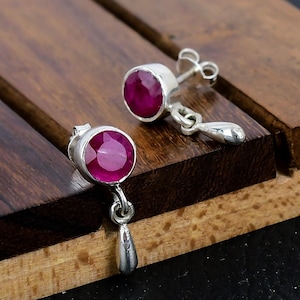 May include: A pair of silver earrings with a dangling teardrop design. Each earring features a round, pink ruby gemstone.