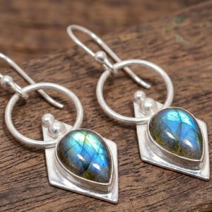 Könnte beinhalten: Silberne Ohrringe mit tropfenförmigen, schillernden blau-grünen Edelsteinen. Die Edelsteine sind in einem rautenförmigen Silberrahmen gefasst, mit einem kreisförmigen Bügel und Hakenverschluss. Auf Holz präsentiert.