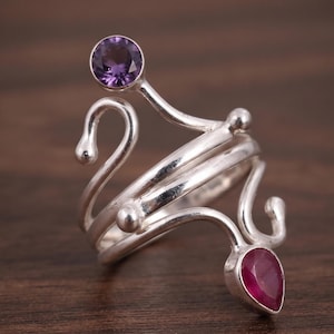 May include: A silver ring featuring a bypass design with a round purple amethyst stone at the top and a pear-shaped red ruby stone at the bottom. The band consists of multiple swirling silver wires that curve into rounded ends, creating an open structure.