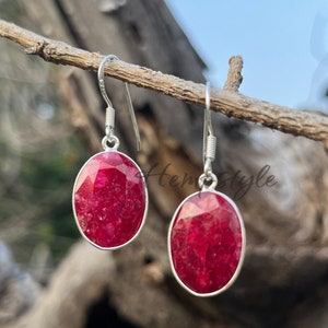 May include: A pair of silver dangle earrings with oval-shaped red ruby gemstones. The earrings are hanging from a branch.