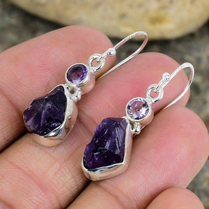 May include: A pair of silver dangle earrings with amethyst gemstones. The earrings feature a larger, rough-cut amethyst stone and a smaller, round amethyst stone.