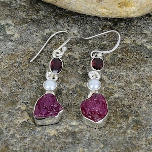 May include: A pair of silver dangle earrings with a red ruby and white pearl accents. The earrings feature a rough, natural ruby gemstone.