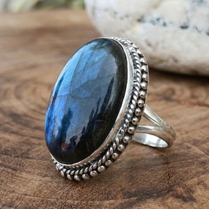 Op de afbeelding: Een zilveren ring met een grote, ovale, blauwe edelsteen met een donker interieur. De edelsteen is gezet in een zilveren band met een kralenrand. De ring wordt gepresenteerd op een houten oppervlak.