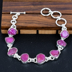 Op de afbeelding: Een zilveren armband met een schuifgesp heeft zeven roze robijn edelstenen. De armband heeft een kettinglink ontwerp en is versierd met zilveren ringen.