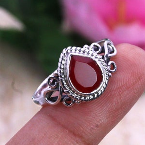 Op de afbeelding: Een zilveren ring met een rode edelsteen in peer-vorm. De ring heeft een delicaat, versierd ontwerp.