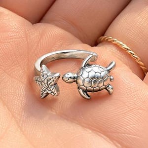 May include: A silver-toned open ring featuring a small sea turtle on one end and a textured starfish on the other. The metal has an oxidized finish to highlight the detailed shell patterns and the star shape, creating a distinct nautical design.